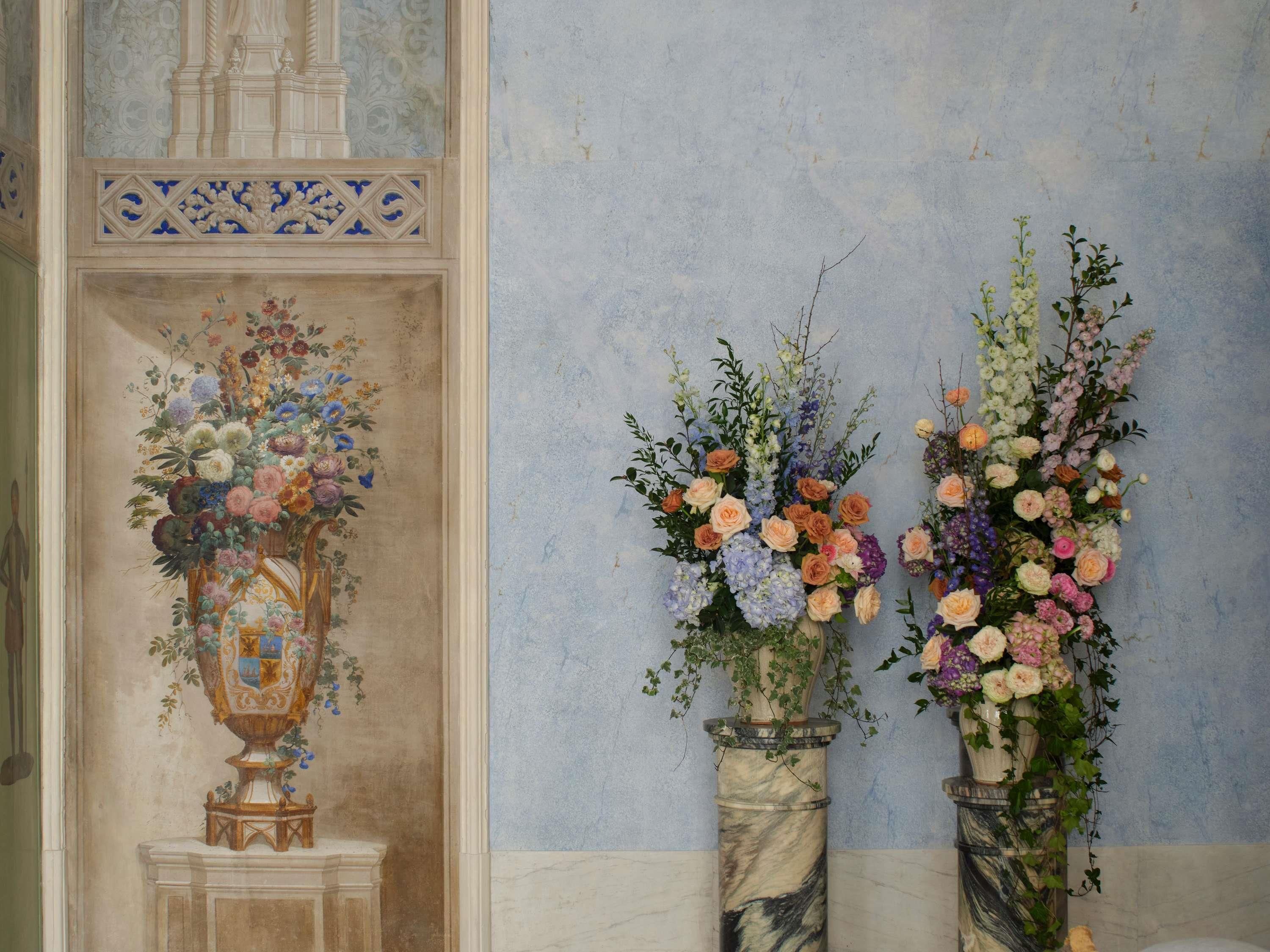A room with a decorative vase on a pedestal, a wall with a floral arrangement, and a marble column.