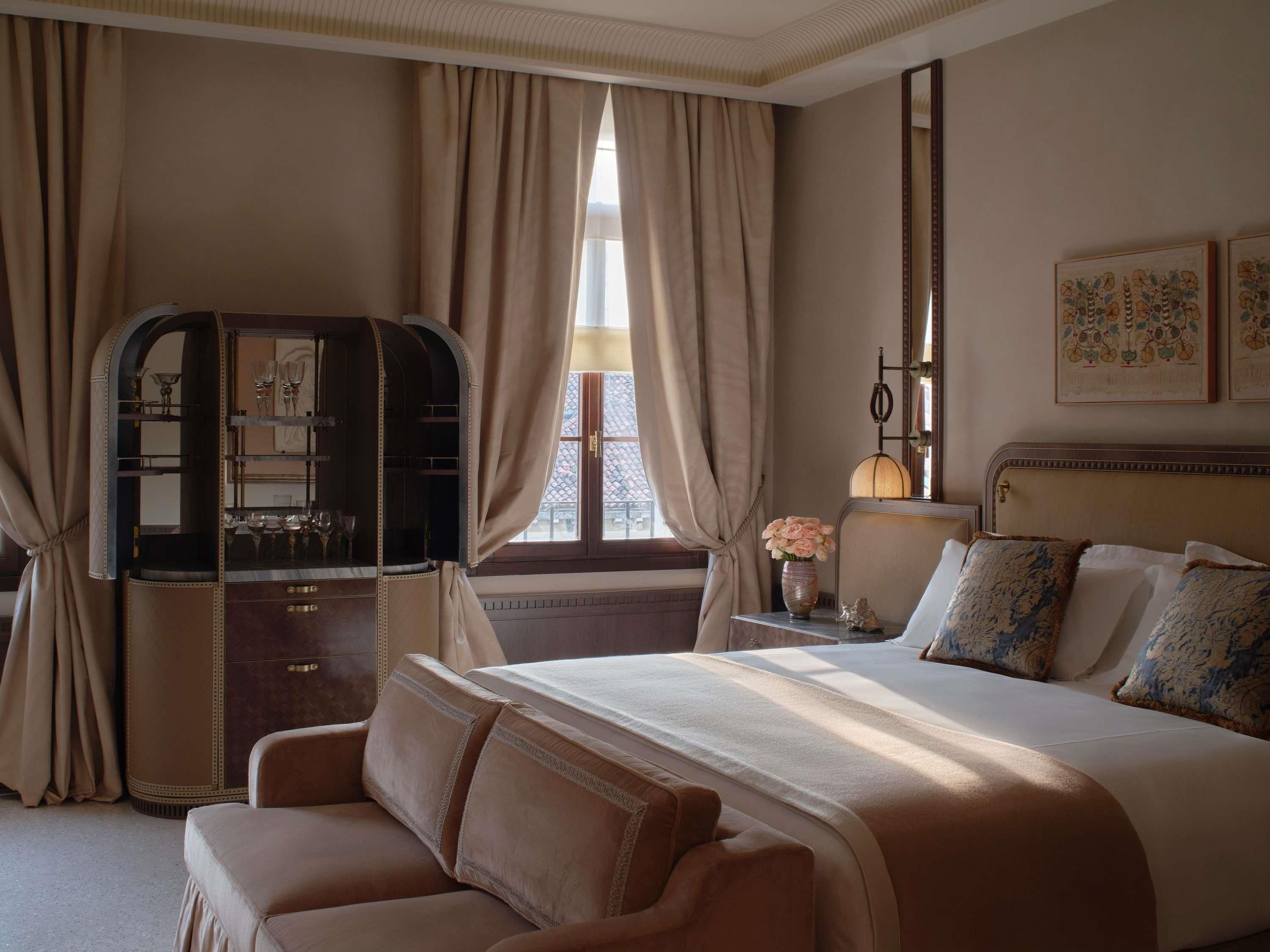 Une chambre d’hôtel comprenant un lit, un canapé, une coiffeuse avec un miroir, une fenêtre avec des rideaux, une table de chevet avec une lampe et des œuvres d’art accrochées au mur.
