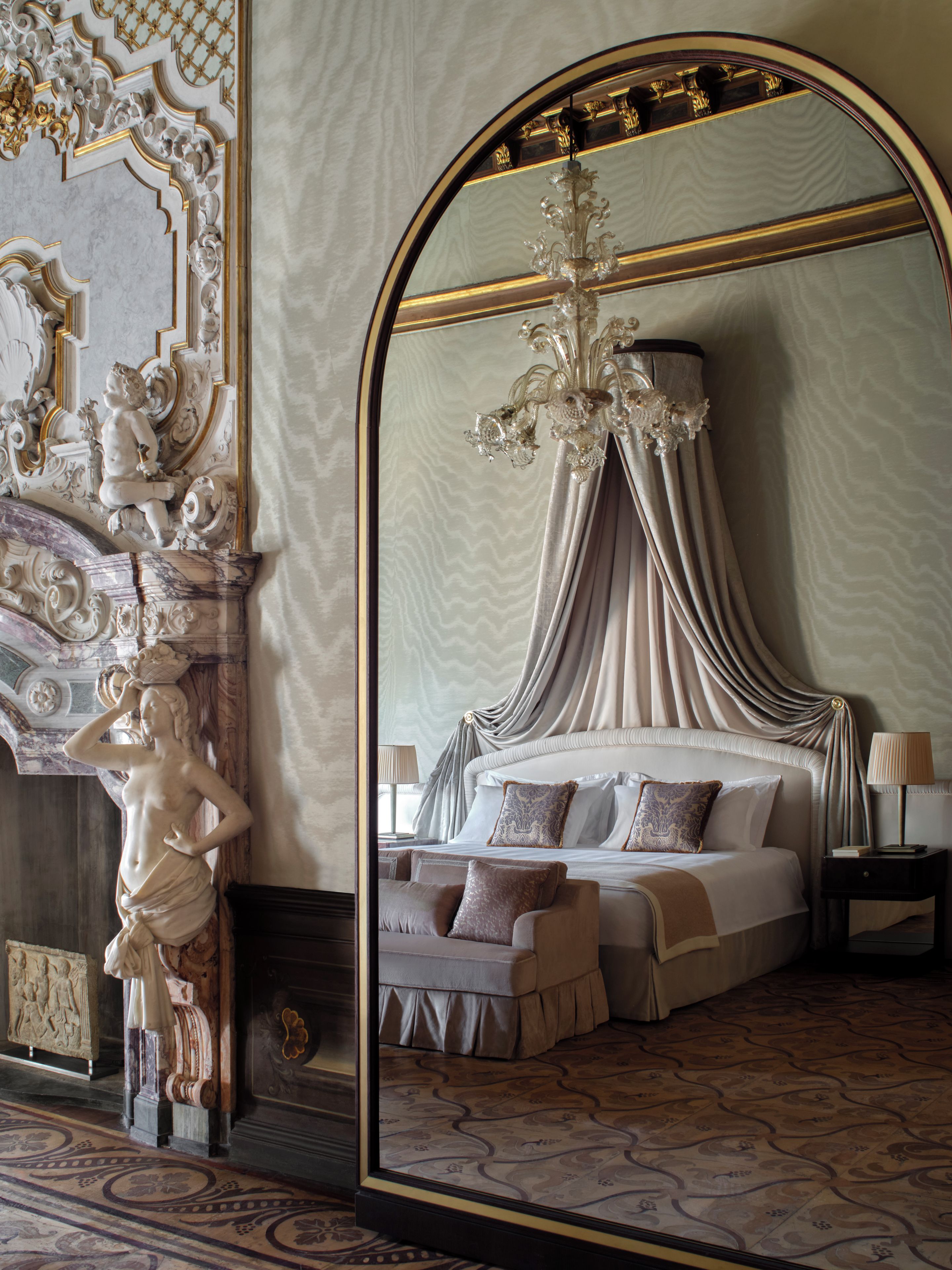 Un dormitorio lujoso con un espejo grande que refleja una cama con cortinas drapeadas, un candelabro y una mesita de noche con una lámpara.