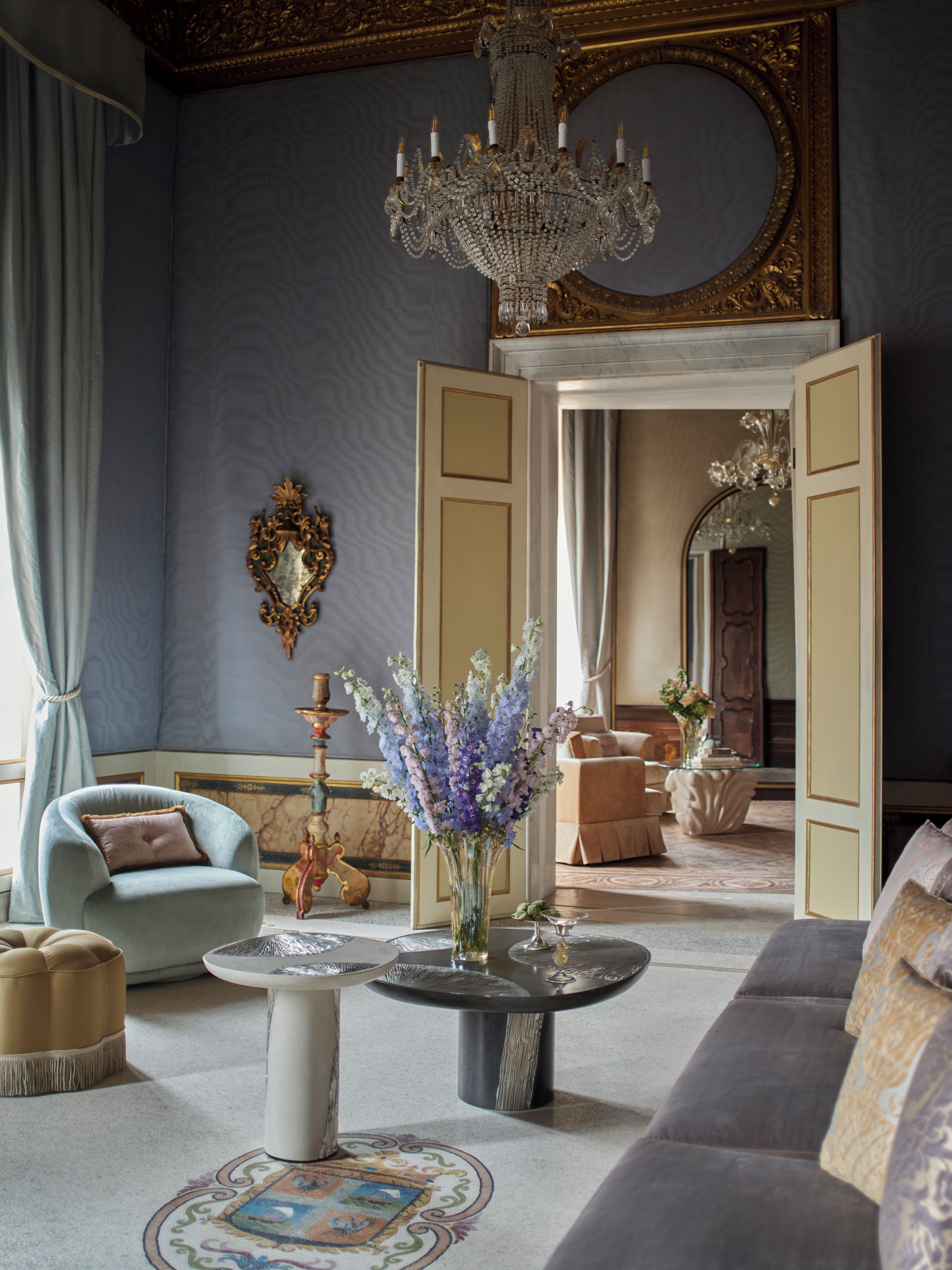 Una habitación lujosa con candelabro, sofá, sillón, mesa de centro con un jarrón de flores y espejo.