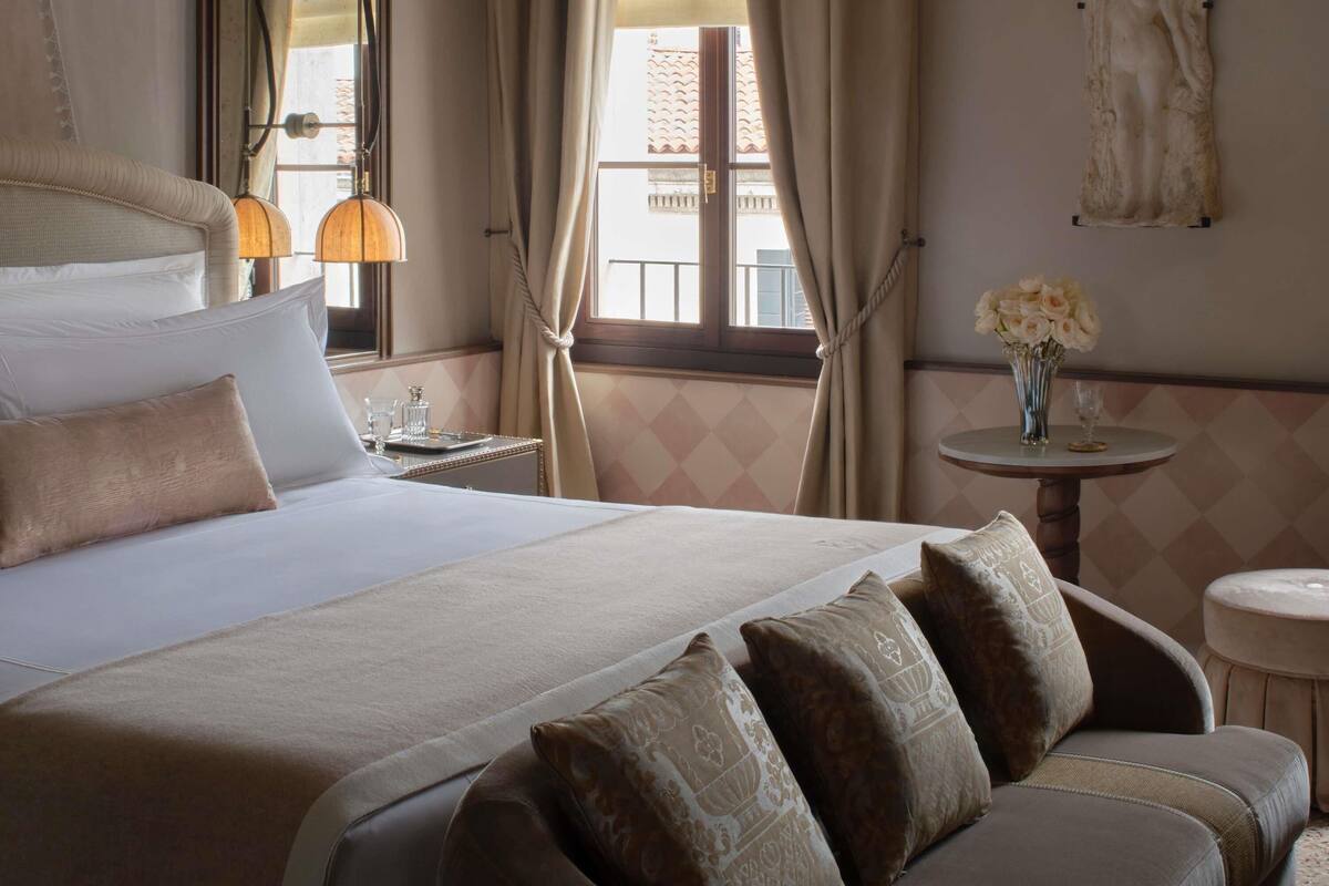 A hotel room with a large bed, a round table with a vase of flowers, and a view of buildings through the window.
