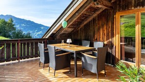 Een houten terras met een tafel en stoelen, een glazen deur naar een balkon, en uitzicht op bergen.