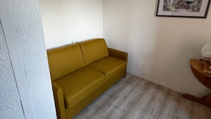 Une petite pièce au mobilier minimaliste, avec un canapé de couleur moutarde, un plancher en bois et un tableau encadré accroché au mur.