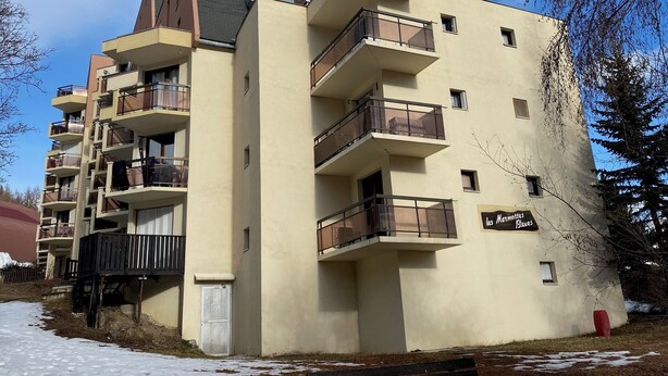 Un immeuble résidentiel à plusieurs étages, doté de balcons, d’une entrée bien visible et d’une enseigne indiquant la présence d’un commerce au rez-de-chaussée.