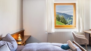 Quarto com cama, escrivaninha, cadeira, abajur e janela com vista para colinas verdes.