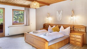 A wooden-framed bedroom with a large bed, bedside tables, a hanging light fixture, and a view of a green landscape through a window.