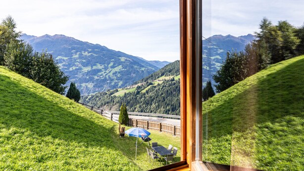 A view from a window overlooking a green hillside with a table and chairs, a blue umbrella, and a distant mountain range. - Double Room 'Zirben Nest' with Stunning View and Private Floor Bathroom (Hainzenberg)
