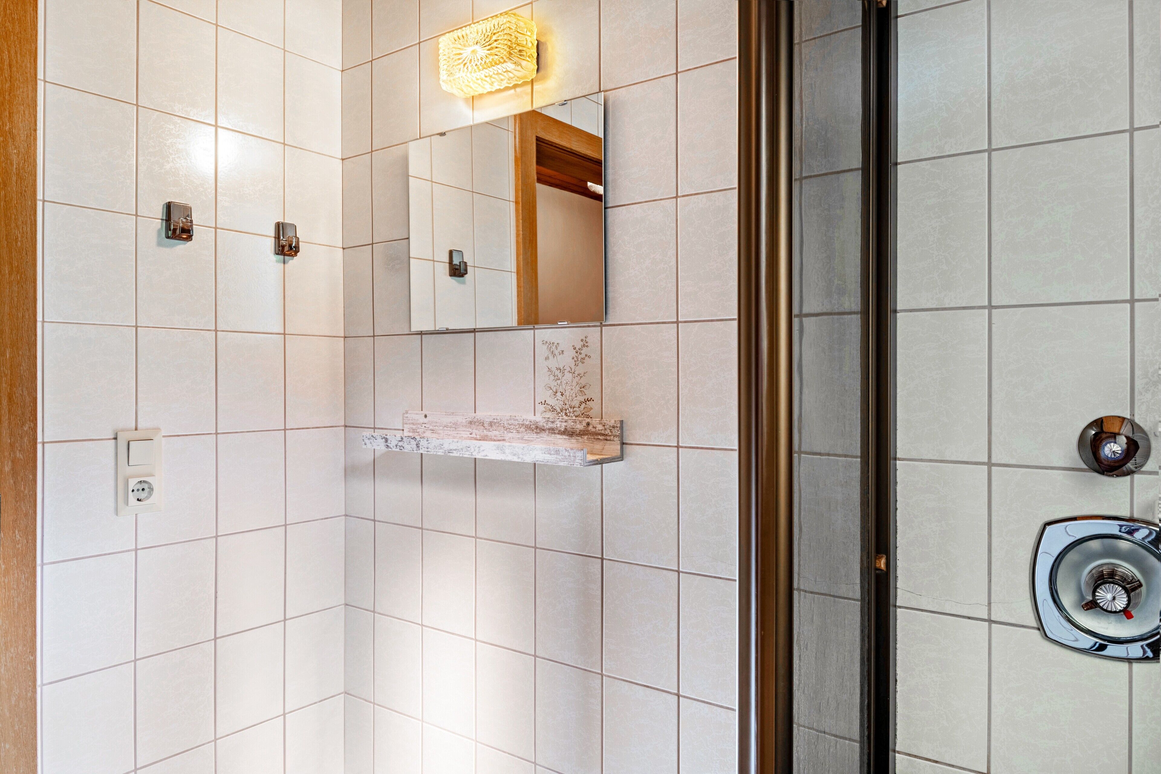 A bathroom with white tiles, a mirror, a shelf, and a shower area.
