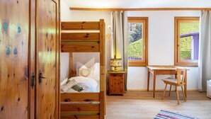 Un lit superposé en bois, un bureau, une chaise et une fenêtre donnant sur un paysage verdoyant.