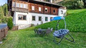 Une maison en bois avec un balcon, un fauteuil suspendu et une table avec des chaises dans la cour.