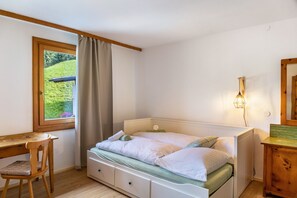 Ein Schlafzimmer mit einem Bett, einem Schreibtisch, einem Stuhl, einem Fenster mit Vorhängen, einer Nachttischlampe und einer Heizung.
