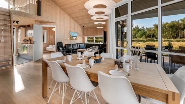 A modern kitchen with white cabinets and a wooden countertop, a dining area with a wooden table and white chairs, and large windows offering a view of the outdoors. - "Kanerva" -  from the sea by Interhome (Ulfborg)
