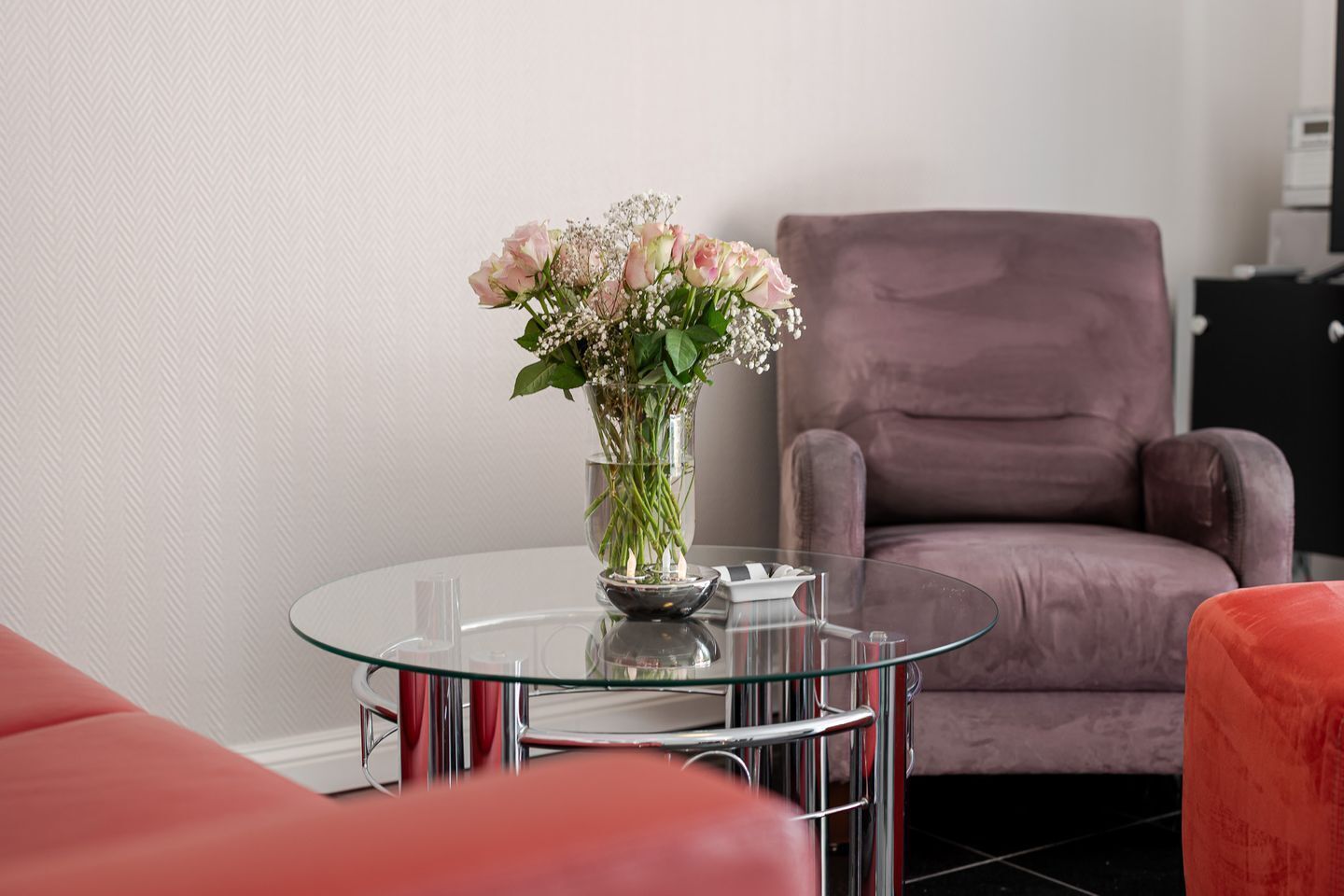 A modern living room with a glass coffee table, a vase of flowers, and a plush armchair.