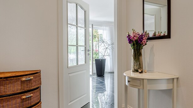Un pasillo con una puerta blanca que da a un balcón, un aparador de mimbre y una consola blanca con un florero.