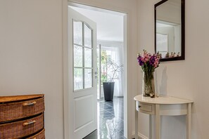 A hallway with a white door leading to a balcony, a wicker chest of drawers, and a white console table with a vase of flowers. - Muschel - Envie de Sylt - Muschel Apartment for 2 people (Sylt-Ost)