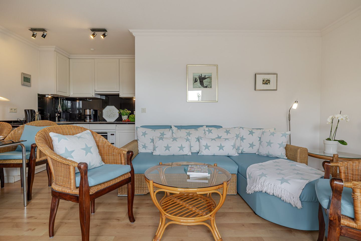 A living room with a blue sofa, wicker chairs, a glass coffee table, and a kitchen in the background.