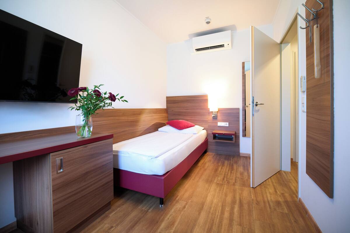 A modern hotel room with a wooden floor, a bed with a red headboard, a bedside table with a lamp, a flat-screen TV mounted on the wall, and an air conditioning unit.