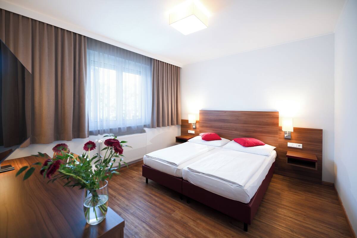 A hotel room with a double bed, a bedside table, a lamp, a wooden headboard, a window with curtains, and a vase with flowers on a wooden desk.