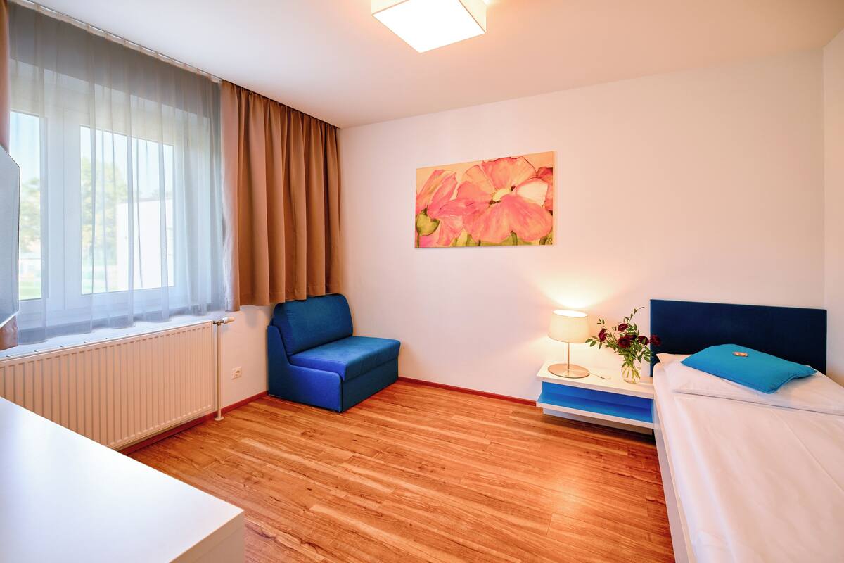 A hotel room with a wooden floor, a blue sofa, a bed with a blue headboard, a nightstand with a lamp, and a painting of flowers on the wall.