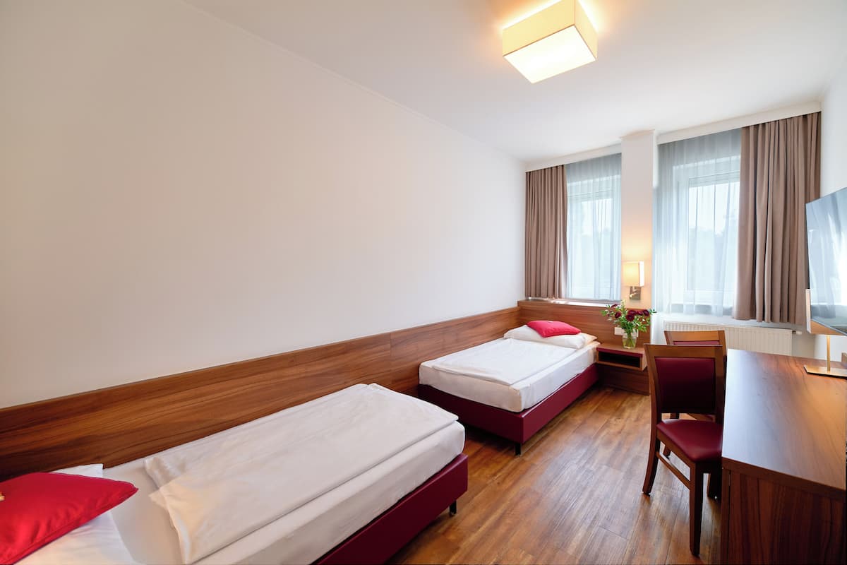A hotel room with two single beds, a wooden headboard, a desk with a chair, a television, and a window with curtains.