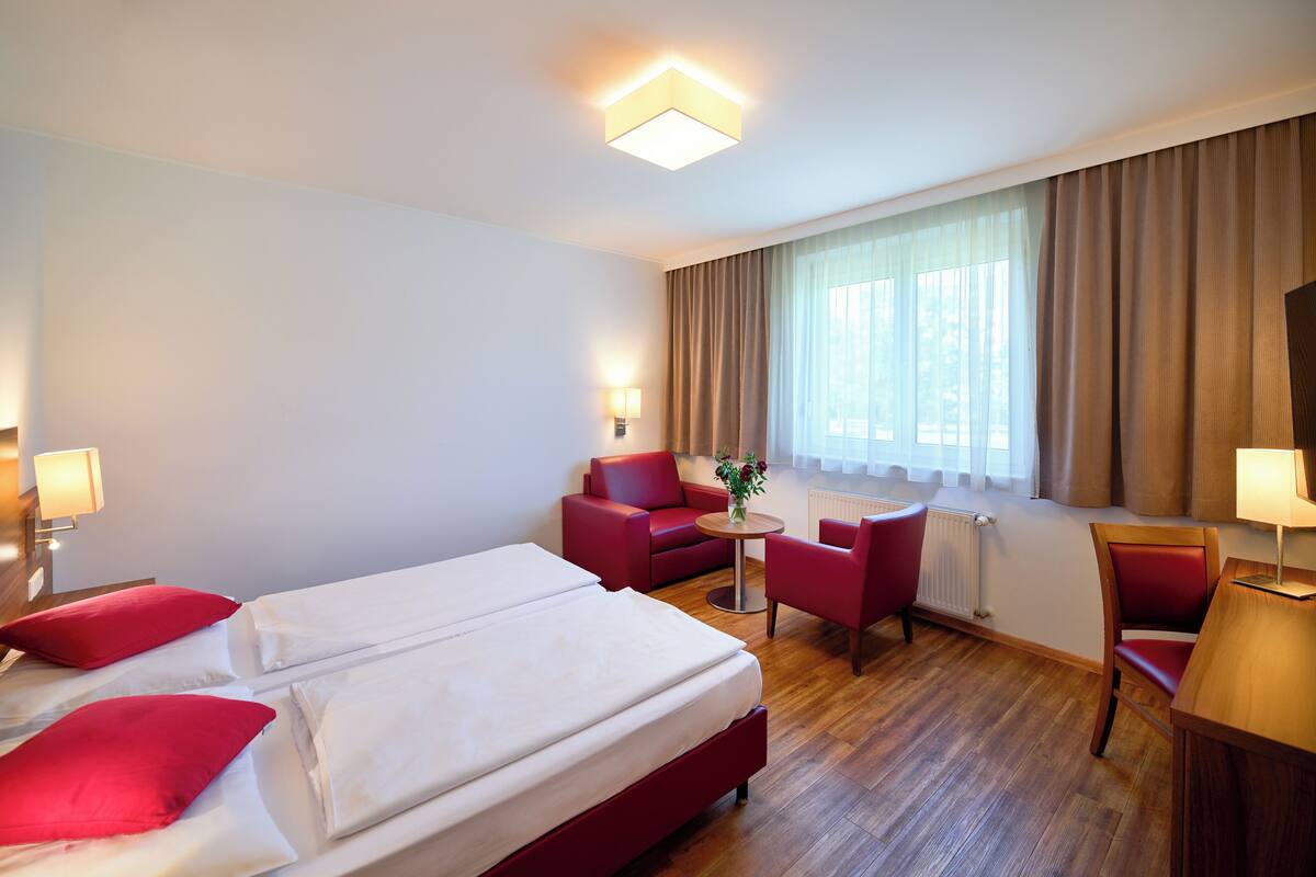 A hotel room with a large bed, two red chairs, a small table with a vase, and a desk with a lamp.