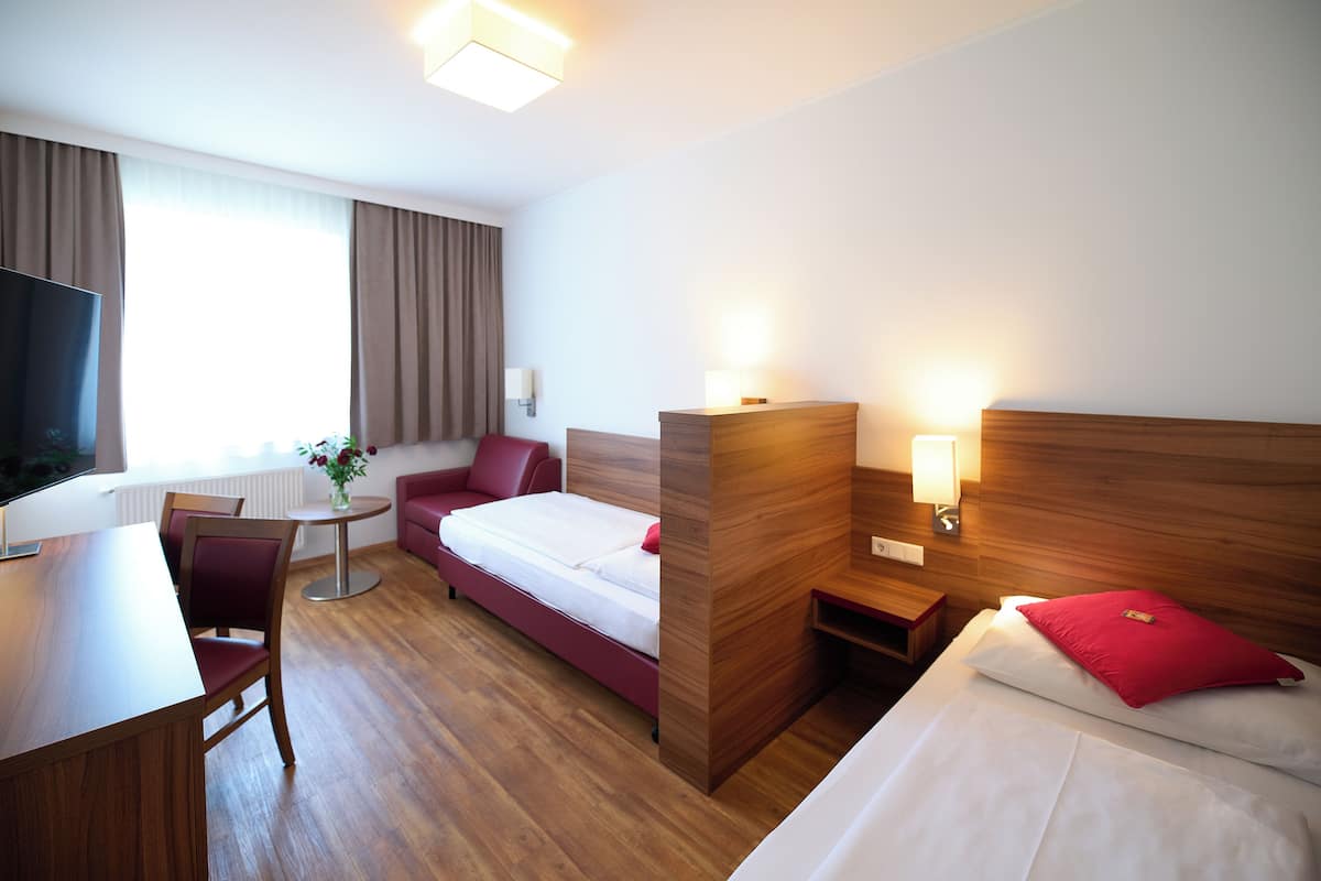 A hotel room with a wooden floor, a bed with white bedding and a red pillow, a red sofa, a small table with a vase of flowers, and a TV on a wooden stand.