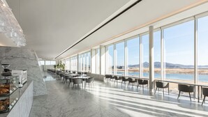Amplio espacio interior con una larga barra mostrando productos de panadería, grandes ventanales que ofrecen vista a una playa y montañas, y un diseño moderno y minimalista.