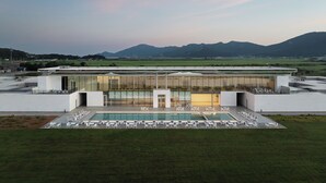 Un edificio moderno con fachada de vidrio, un área de piscina con sillas blancas y vistas a montañas distantes.