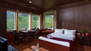 A hotel room with a bed, a desk, and a chair. There are windows with a view of greenery outside.