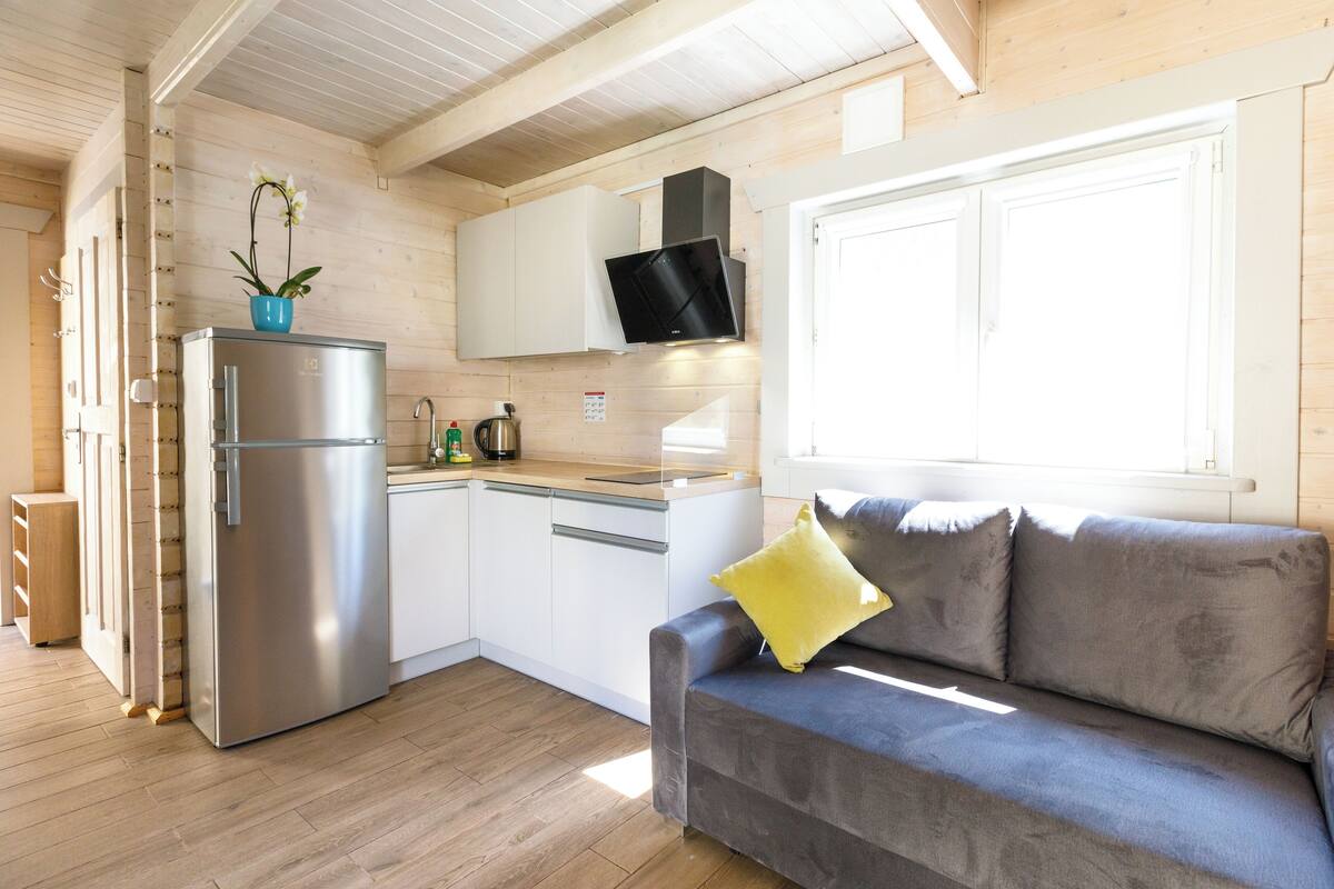 Una cocina compacta con refrigerador integrado, microondas y estufa. Hay un sofá gris con un cojín amarillo y una ventana con persianas.