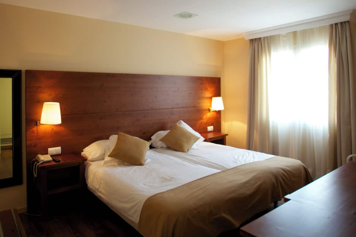 A hotel room with a large bed, two bedside tables with lamps, a wooden headboard, and a window with curtains.
