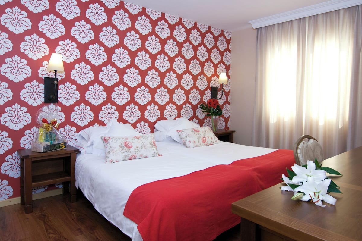 A hotel room with a bed, a wooden nightstand, a table with a vase of flowers, and a patterned wallpaper.