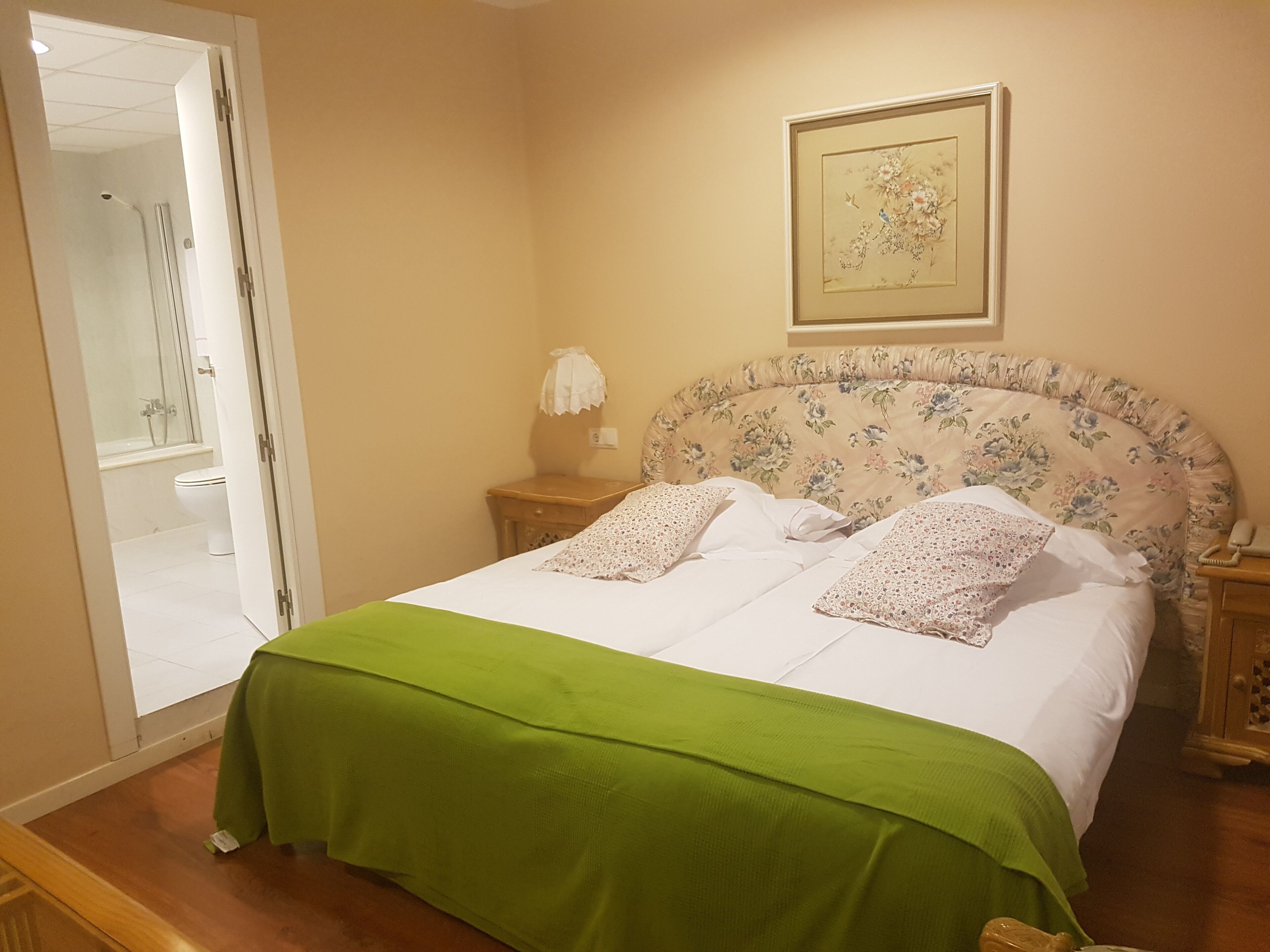 Ein ordentlich bezogenes Bett mit grüner Bettdecke und Kissen mit Blumenmuster, ein Nachttisch aus Holz mit einer Lampe und ein Blumenbild in einem Rahmen an der Wand.