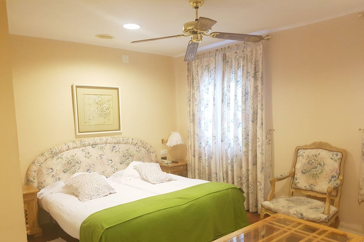 A bedroom with a bed, a green bedspread, a ceiling fan, a chair, and a framed picture on the wall.
