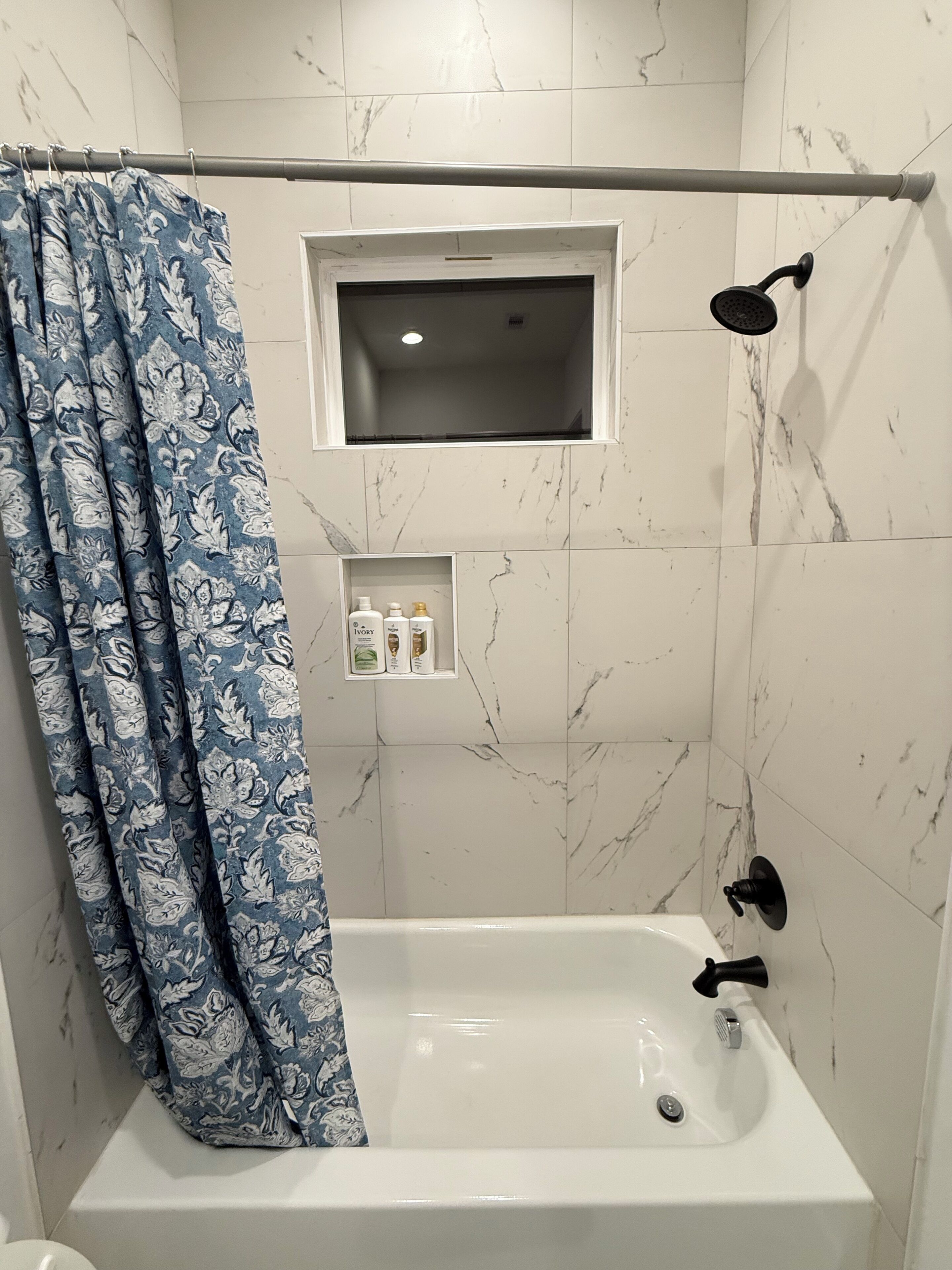 Un baño con bañera, ducha, una ventana y un estante con artículos de aseo.