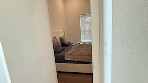 Un couloir étroit mène à une chambre à coucher équipée d’un lit, d’un ventilateur de plafond et d’une fenêtre avec des stores.