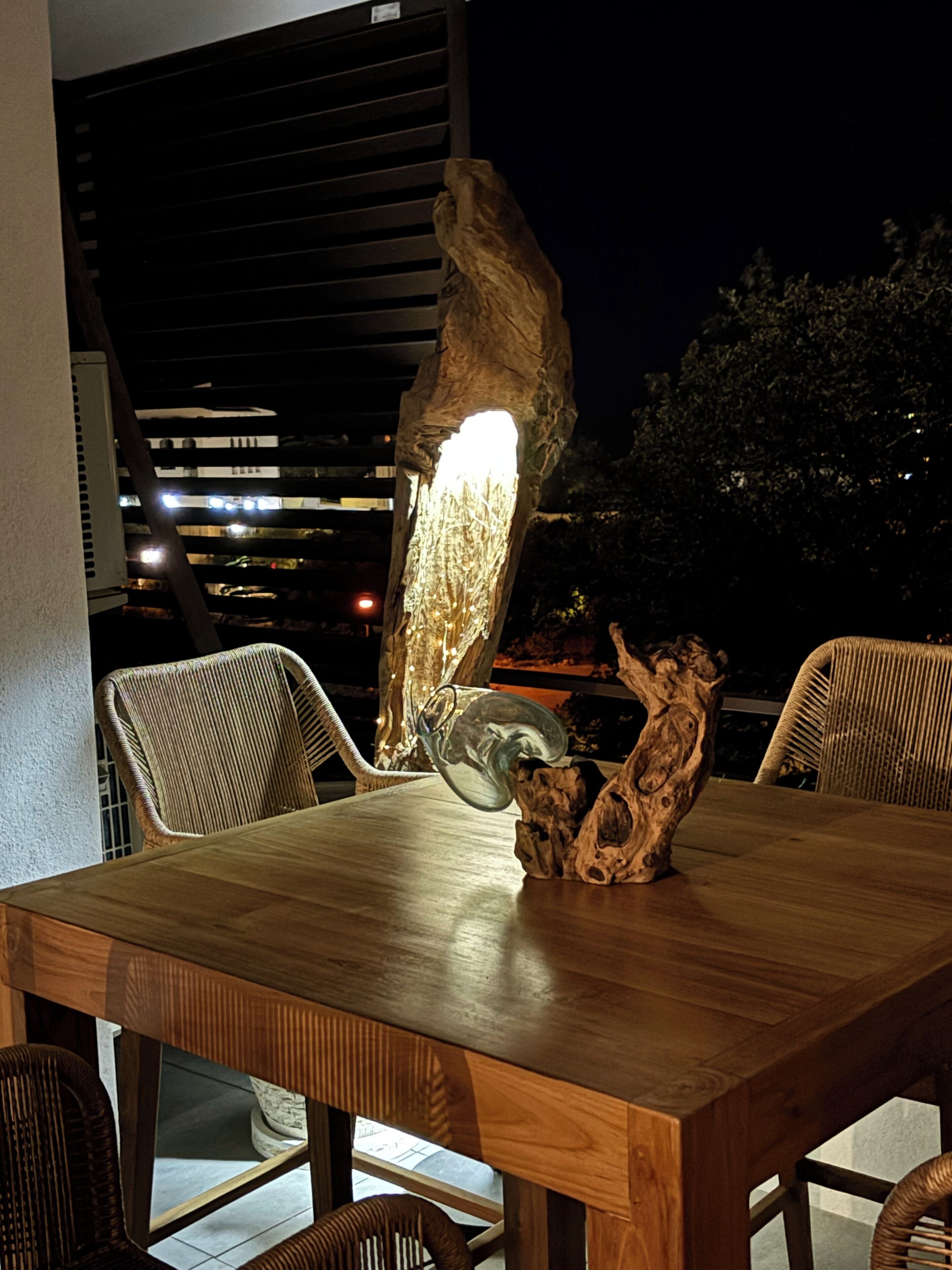 An outdoor dining area with a wooden table and wicker chairs, featuring a unique light fixture and a sculptural centerpiece.