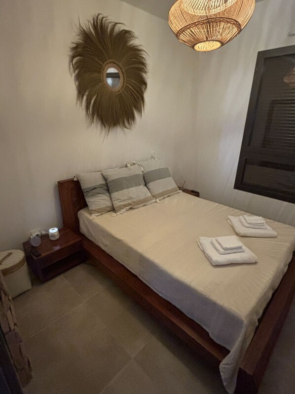 A neatly made bed with a white comforter and pillows, a bedside table with a lamp, and a wall-mounted mirror.