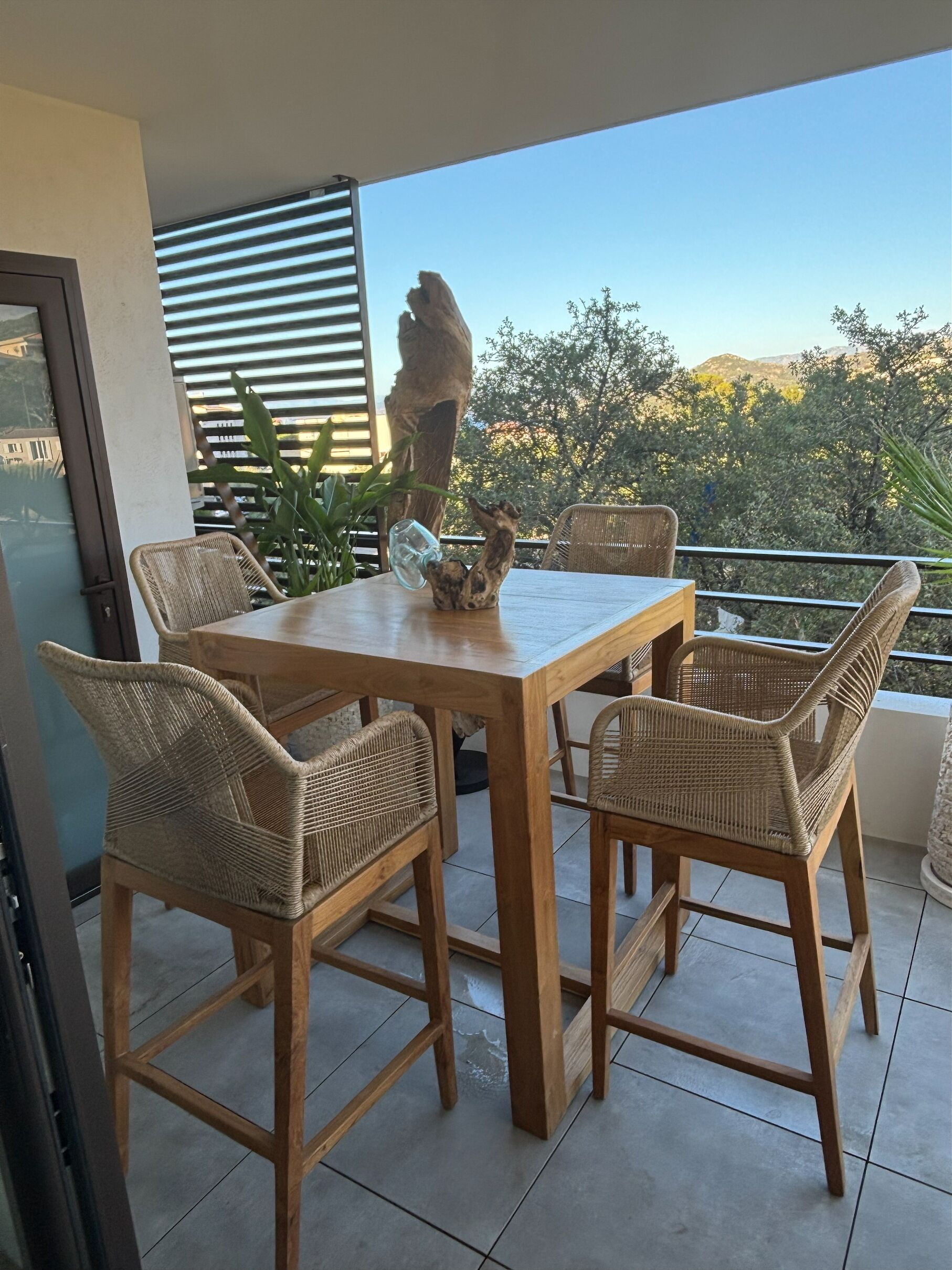 A wooden table with four wicker chairs on a balcony with a view of trees and hills.