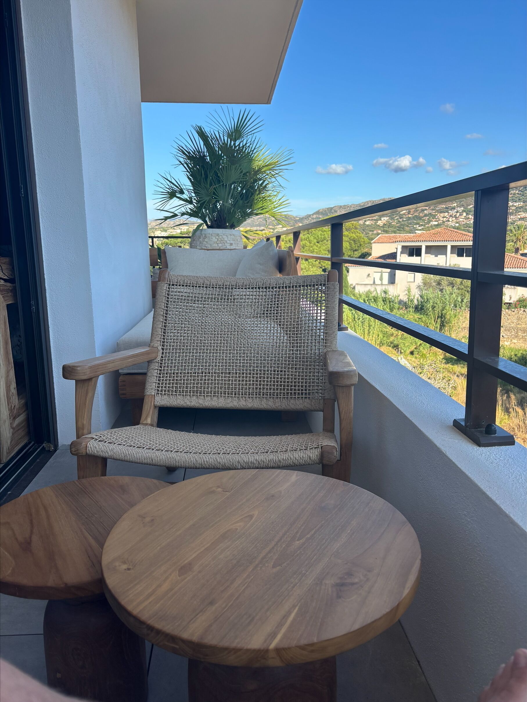 Een balkon met een rieten stoel en een ronde houten tafel, met uitzicht op een woonwijk met huizen en groen.