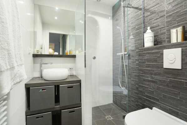 A modern bathroom with a glass-enclosed shower, a white sink, and a dark tiled wall. - Chic Apartment & Terrace Eiffel Tower - Trocadero (Paris)