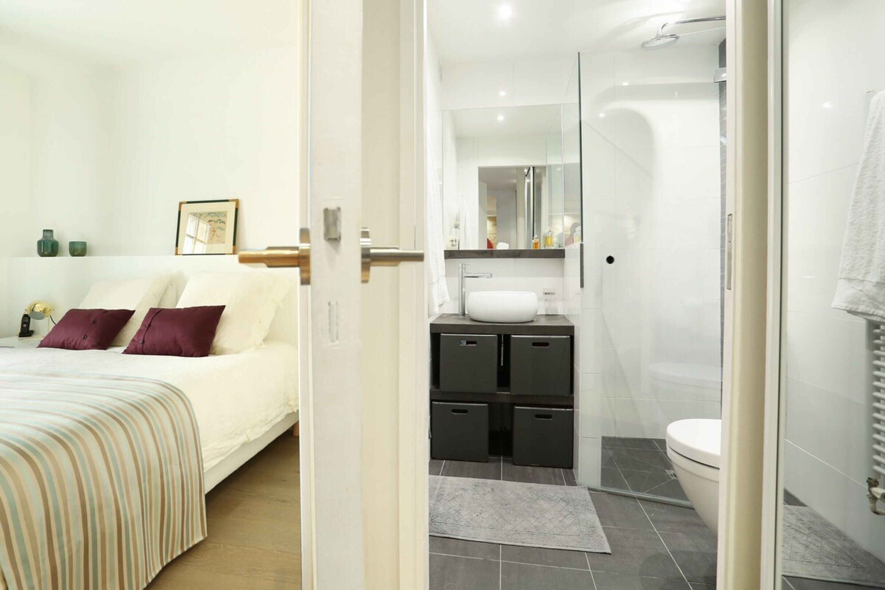 A modern bedroom with a bed, bedside tables, and a nightstand. Adjacent to the bedroom is a bathroom with a shower, sink, and toilet.