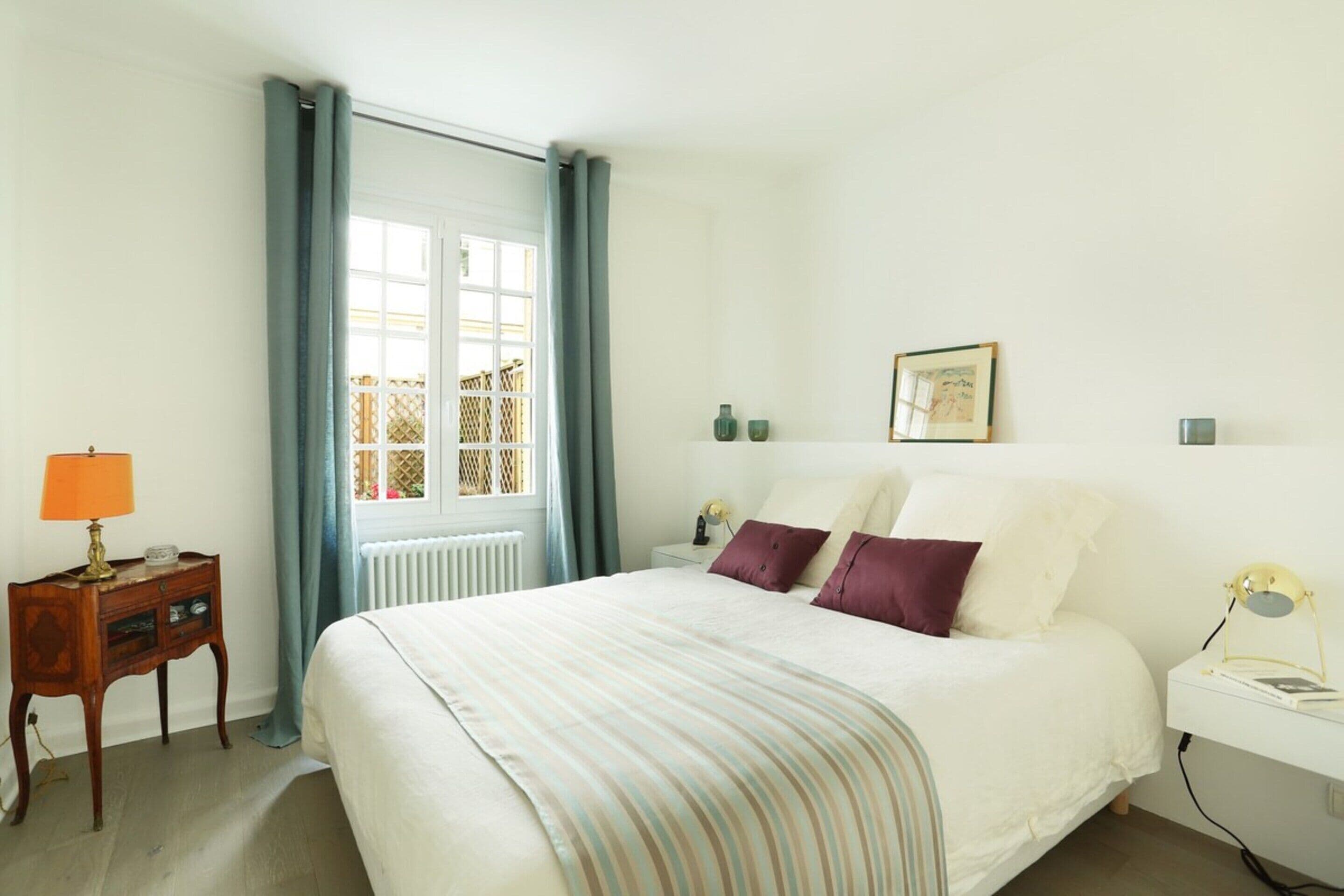 A bedroom with a bed, a nightstand, a lamp, a window with curtains, and a wall-mounted picture.