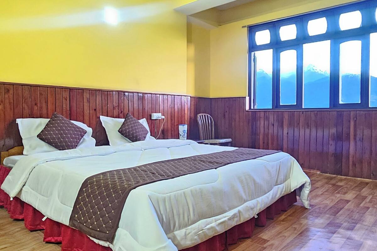 A hotel room with a wooden headboard, a large bed with multiple pillows, a window with a view of mountains, and a wooden floor.