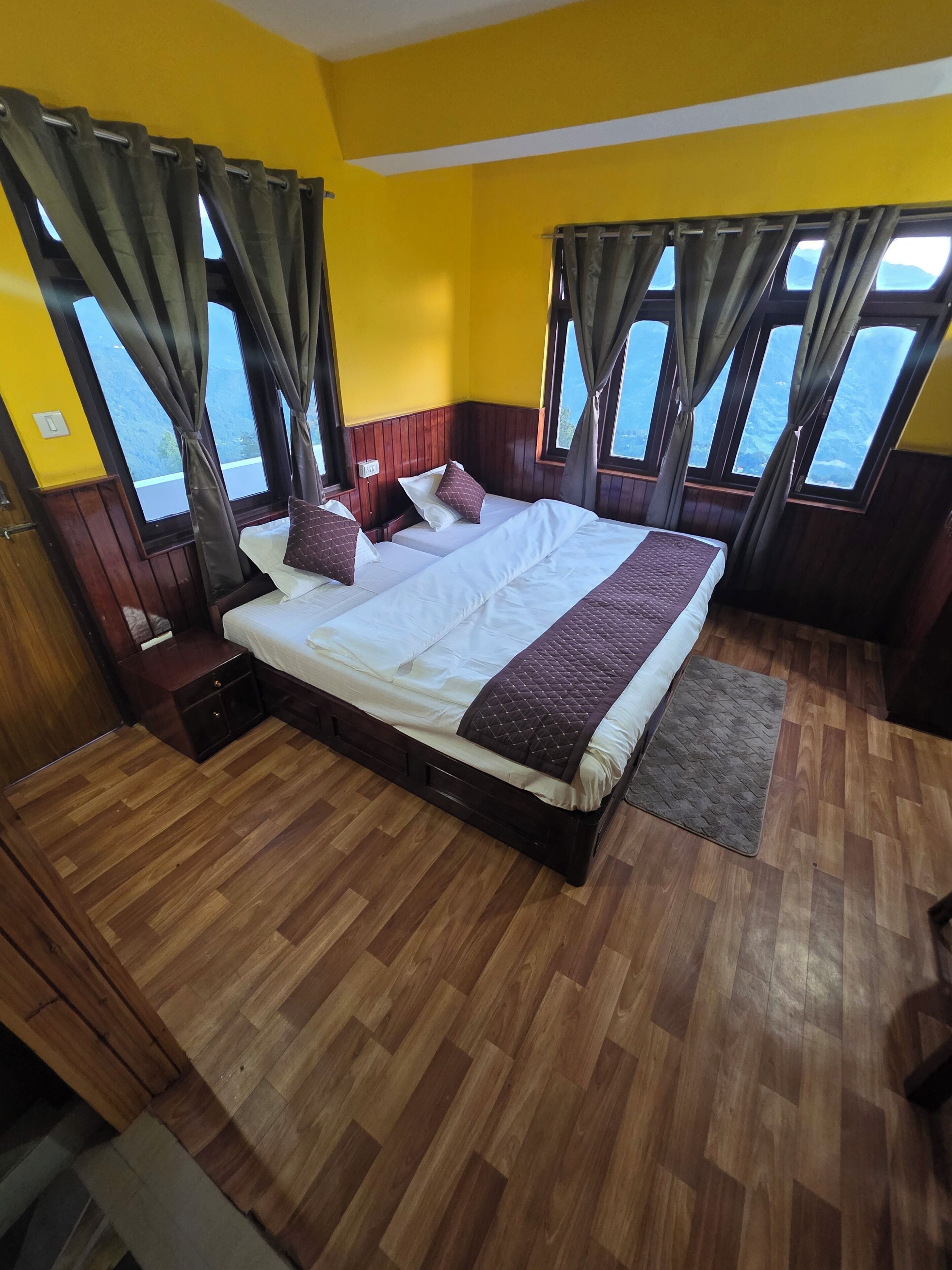 Un dormitorio con una cama grande, suelo de madera y cortinas sobre las ventanas.
