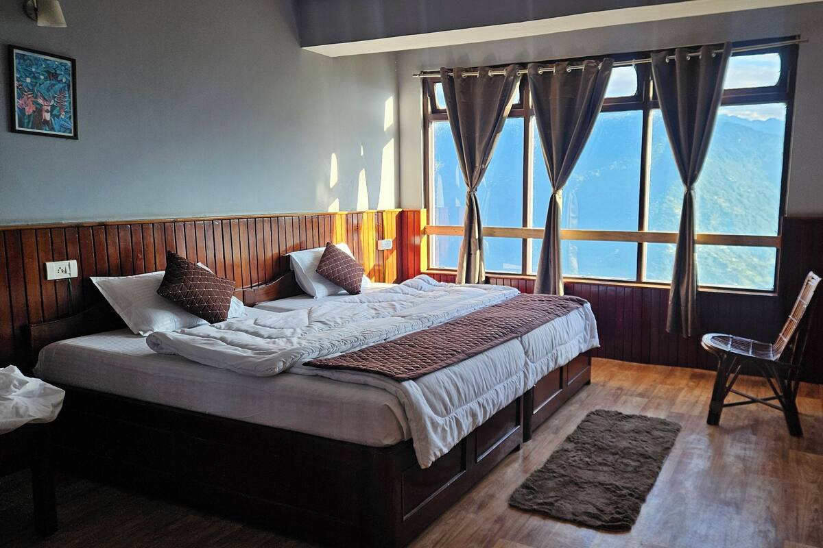 A bedroom with a wooden headboard, a bed with white and brown bedding, a chair, and a view of the outdoors through large windows.