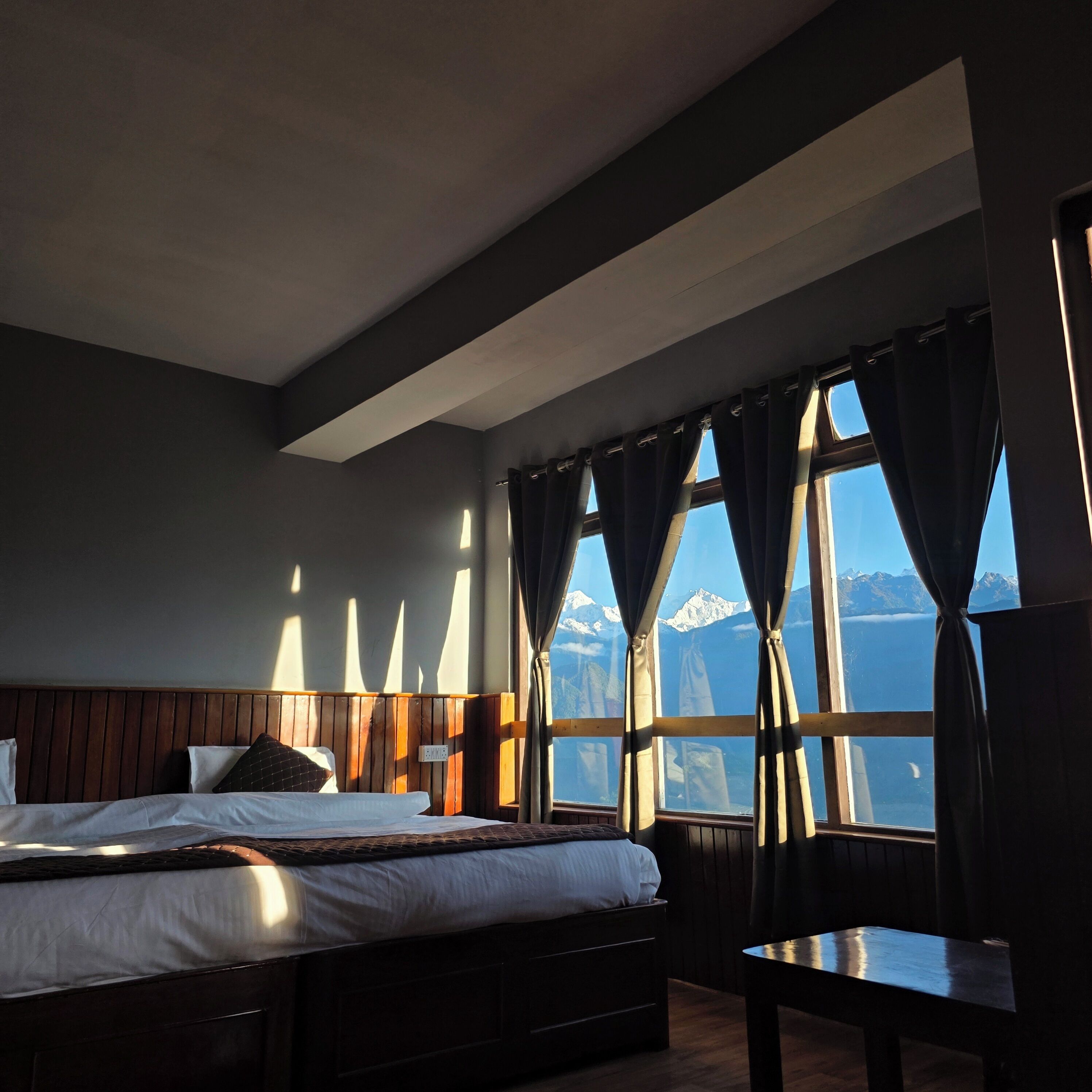 A hotel room with a bed, a wooden headboard, a window with curtains, and a view of mountains.