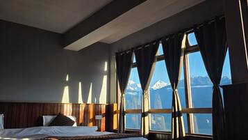 A hotel room with a bed, a wooden headboard, a window with curtains, and a view of mountains.