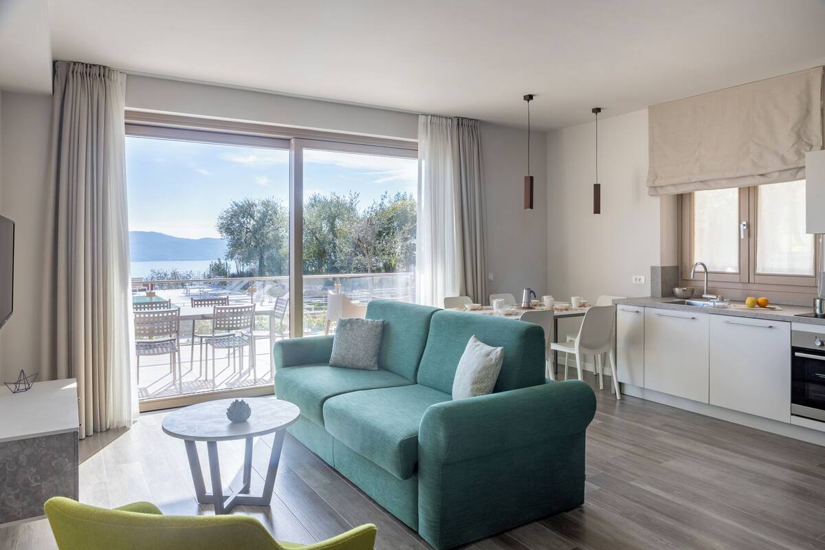 A modern living room with a green sofa, a small round table, and a view of a balcony with outdoor furniture.