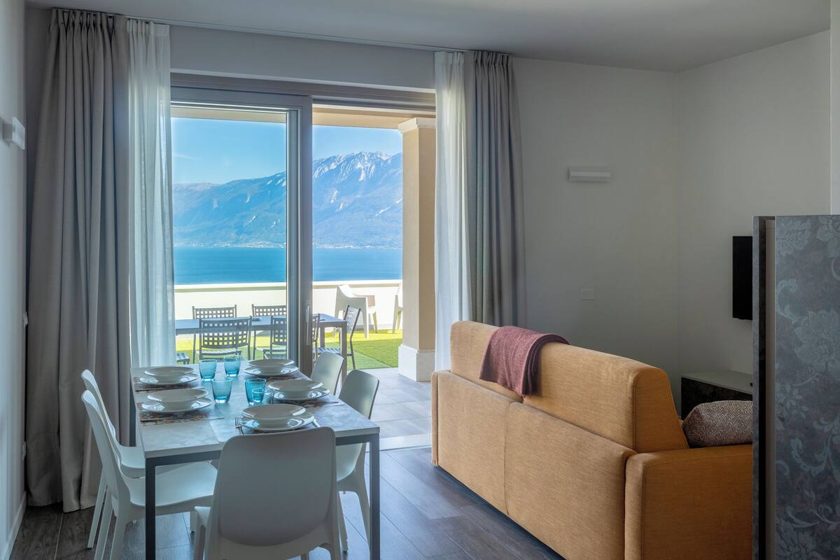 A modern living room with a sofa and a dining table set for four, a TV, and a view of a mountainous landscape through large windows.